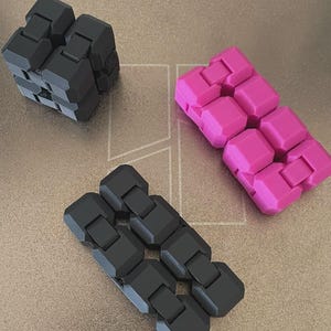 以下が含まれることがあります： プラスチック製の関節式フィジェットトイ3点。黒い立方体型が1つと、黒と鮮やかなピンクの長方形型が2つ。各おもちゃは複数の連結されたセグメントで構成されており、折りたたんだり動かしたりして遊ぶことができる構造になっています。