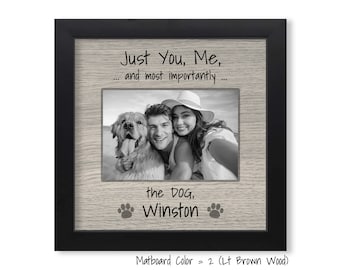 May include: A black picture frame with a light brown wood-grain mat. The frame has a photo of a couple and their dog. The text "Just You, Me, and most importantly... the DOG, Winston" is written on the mat.