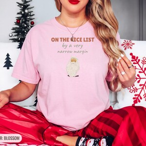 Blossom Comfort Colors t-shirt printed with the words On The Nice List By A Very Narrow Margin and a drawing of a red santa hat beside a candy cane, worn by a woman