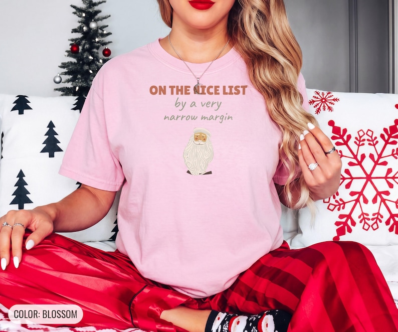 Blossom Comfort Colors t-shirt printed with the words On The Nice List By A Very Narrow Margin and a drawing of a red santa hat beside a candy cane, worn by a woman