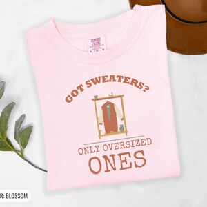 Blossom Comfort Colors t-shirt printed with the words Got Sweaters? Only Oversized Ones and a drawing of a knitted wool cardigan hanging from a simple wooden clothes hanger, folded flat lay