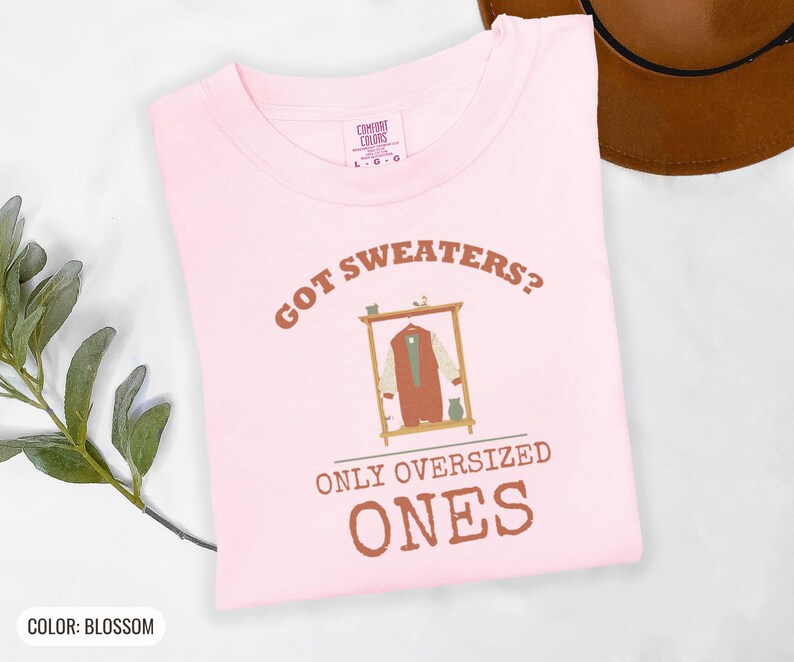 Blossom Comfort Colors t-shirt printed with the words Got Sweaters? Only Oversized Ones and a drawing of a knitted wool cardigan hanging from a simple wooden clothes hanger, folded flat lay