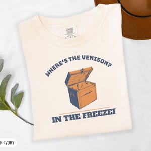Ivory Comfort Colors t-shirt printed with the words Where'S The Venison? In The Freezer and a drawing of a vintage metal chest freezer with a pair of paper-wrapped butcher packages resting on top, folded flat lay
