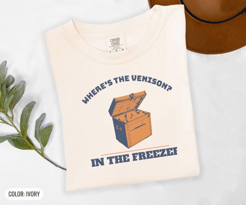 Ivory Comfort Colors t-shirt printed with the words Where'S The Venison? In The Freezer and a drawing of a vintage metal chest freezer with a pair of paper-wrapped butcher packages resting on top, folded flat lay