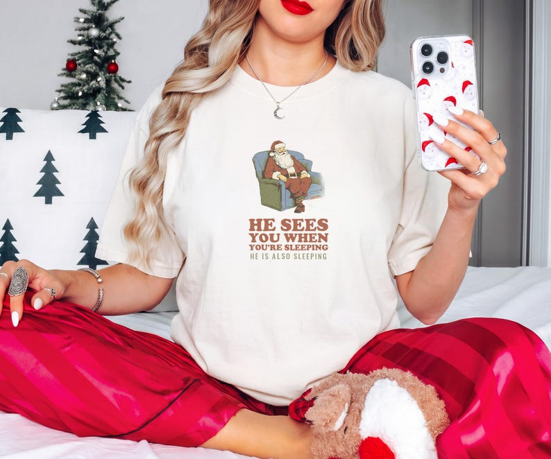 Ivory Comfort Colors t-shirt printed with the words He Sees You When You'Re Sleeping He Is Also Sleeping and a drawing of santa Claus in unbuttoned flannel asleep on a patterned sofa beside a drowsy elf wearing a, worn by a woman