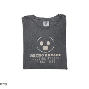 Pepper Comfort Colors t-shirt printed with the words Retro Arcade Dodging Ghosts Since 1980 and a drawing of a maze with a Pac-Man character, folded flat lay