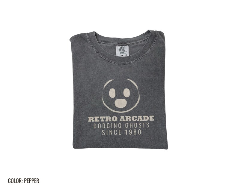 Pepper Comfort Colors t-shirt printed with the words Retro Arcade Dodging Ghosts Since 1980 and a drawing of a maze with a Pac-Man character, folded flat lay