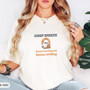 Ivory Comfort Colors t-shirt printed with the words Crisp Breeze Leaves Turning And Bones Rattling and a drawing of a large cute smiling ghost beside a carved jack-o-lantern, worn by a woman