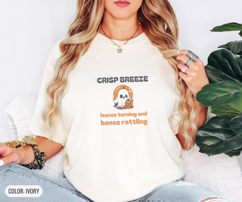 Ivory Comfort Colors t-shirt printed with the words Crisp Breeze Leaves Turning And Bones Rattling and a drawing of a large cute smiling ghost beside a carved jack-o-lantern, worn by a woman