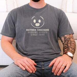 Pepper Comfort Colors t-shirt printed with the words Retro Arcade Dodging Ghosts Since 1980 and a drawing of a maze with a Pac-Man character, worn by a man