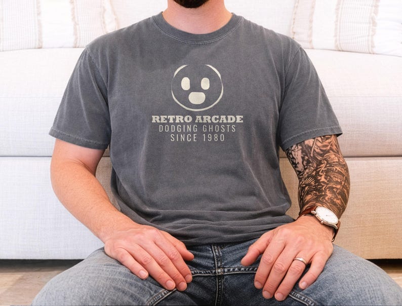 Pepper Comfort Colors t-shirt printed with the words Retro Arcade Dodging Ghosts Since 1980 and a drawing of a maze with a Pac-Man character, worn by a man