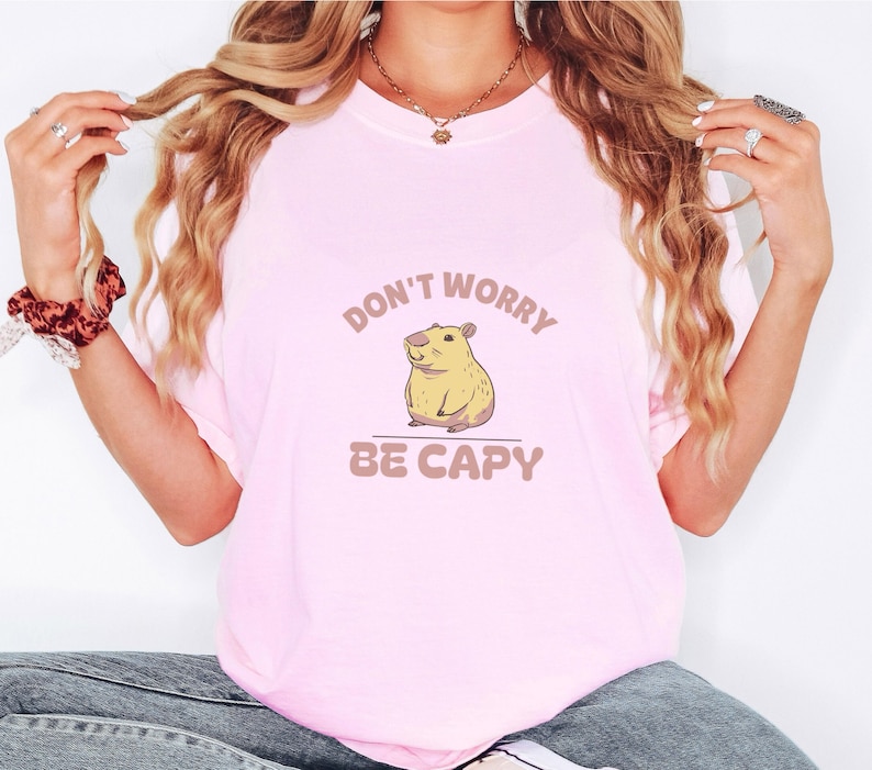 Blossom Comfort Colors t-shirt printed with the words Don'T Worry Be Capy and a drawing of a happy capybara with a small orange citrus slice on its head beside a green lily pad, worn by a woman
