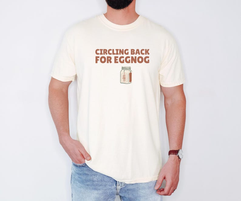 Ivory Comfort Colors t-shirt printed with the words Circling Back For Eggnog and a drawing of a vintage paper milk carton labeled eggnog with a green pine needle design on the side, worn by a man