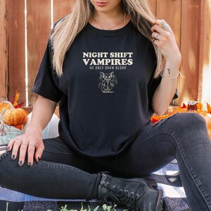 Black Comfort Colors t-shirt printed with the words Night Shift Vampires We Only Draw Blood and a drawing of a cute cartoon vampire bat wearing a tiny nurse scrub top, worn by a woman