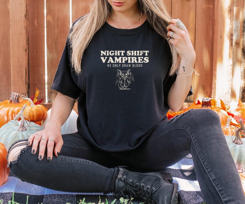 Black Comfort Colors t-shirt printed with the words Night Shift Vampires We Only Draw Blood and a drawing of a cute cartoon vampire bat wearing a tiny nurse scrub top, worn by a woman