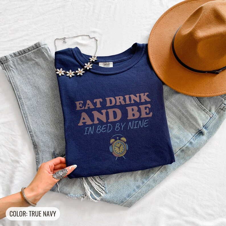 True Navy Comfort Colors t-shirt printed with the words Eat Drink And Be In Bed By Nine and a drawing of a retro brass wind-up alarm clock with its bells wrapped in fuzzy winter earmuffs, folded flat lay