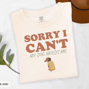 Ivory Comfort Colors t-shirt printed with the words Sorry I Can'T My Dog Needs Me and a drawing of a sleeping dachshund wearing a patterned knit sweater, folded flat lay