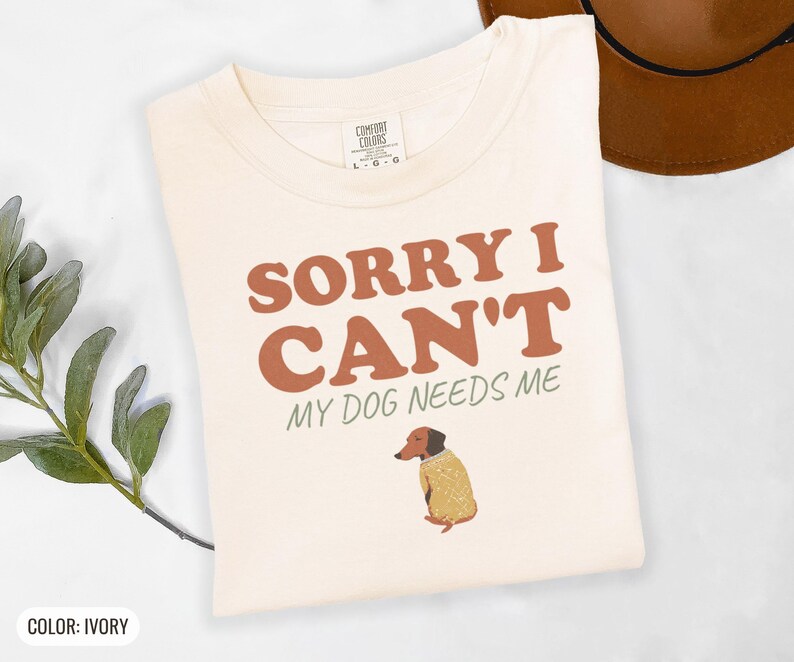 Ivory Comfort Colors t-shirt printed with the words Sorry I Can'T My Dog Needs Me and a drawing of a sleeping dachshund wearing a patterned knit sweater, folded flat lay