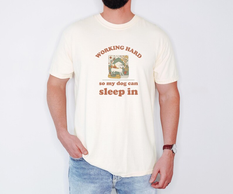 Ivory Comfort Colors t-shirt printed with the words Working Hard So My Dog Can Sleep In and a drawing of a dachshund lounging on a plush pillow with sunglasses on its face, worn by a man