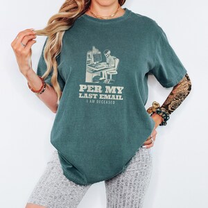 Blue Spruce Comfort Colors t-shirt printed with the words Per My Last Email I Am Deceased and a drawing of a skeleton sitting at an office desk typing on a computer keyboard, worn by a woman