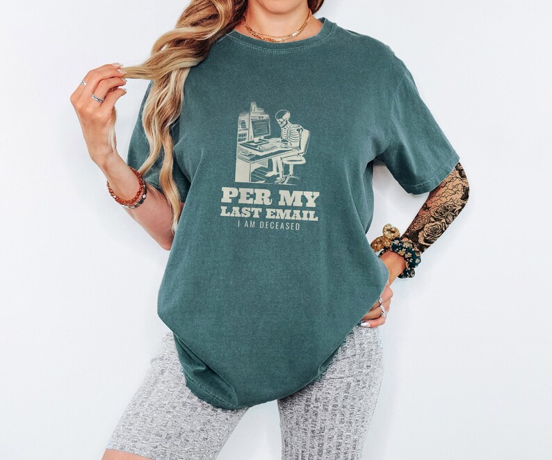 Blue Spruce Comfort Colors t-shirt printed with the words Per My Last Email I Am Deceased and a drawing of a skeleton sitting at an office desk typing on a computer keyboard, worn by a woman