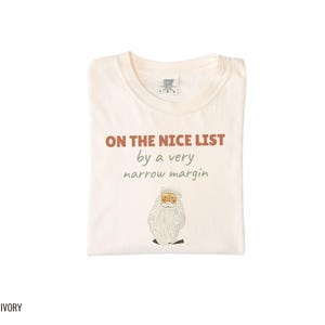 Ivory Comfort Colors t-shirt printed with the words On The Nice List By A Very Narrow Margin and a drawing of a red santa hat beside a candy cane, folded flat lay