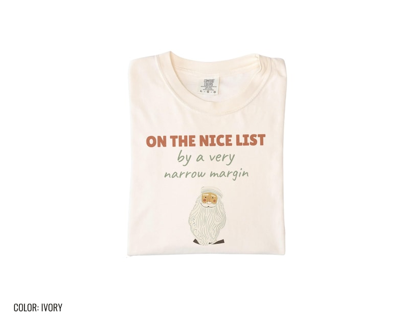 Ivory Comfort Colors t-shirt printed with the words On The Nice List By A Very Narrow Margin and a drawing of a red santa hat beside a candy cane, folded flat lay