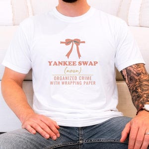 White Comfort Colors t-shirt printed with the words Yankee Swap (Noun) Organized Crime With Wrapping Paper and a drawing of a large red comical bow with long satin ribbon tails, worn by a man
