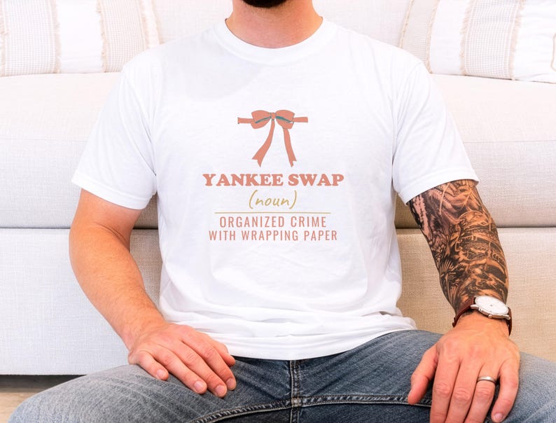 White Comfort Colors t-shirt printed with the words Yankee Swap (Noun) Organized Crime With Wrapping Paper and a drawing of a large red comical bow with long satin ribbon tails, worn by a man
