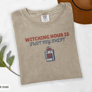 Khaki Comfort Colors t-shirt printed with the words Witching Hour Is Just My Shift and a drawing of a retro medical defibrillator machine decorated with a spooky jack-o-lantern face on its m, folded flat lay