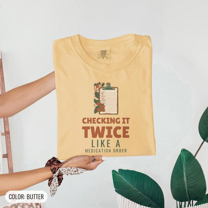 Butter Comfort Colors t-shirt printed with the words Checking It Twice Like A Medication Order and a drawing of a classic wooden medical clipboard holding a paper medication checklist with checkboxes, a, folded flat lay