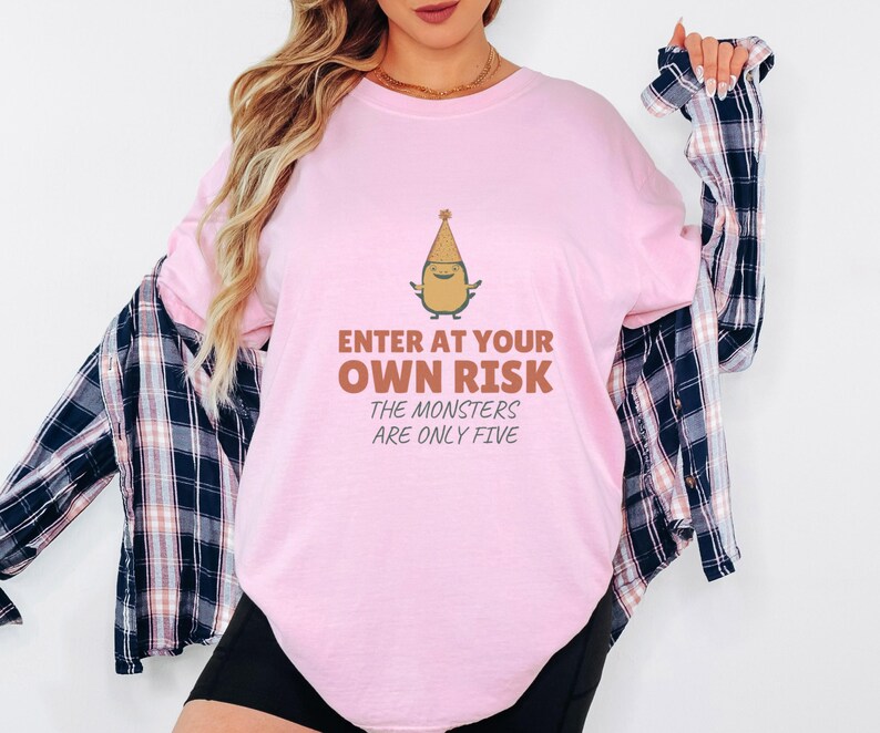 Blossom Comfort Colors t-shirt printed with the words Enter At Your Own Risk The Monsters Are Only Five and a drawing of a cute, smiling little green monster wearing a tiny paper party hat and holding a crayon, worn by a woman