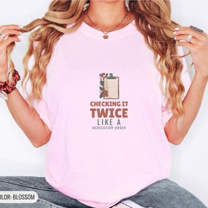 Blossom Comfort Colors t-shirt printed with the words Checking It Twice Like A Medication Order and a drawing of a classic wooden medical clipboard holding a paper medication checklist with checkboxes, a, worn by a woman
