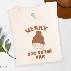 Ivory Comfort Colors t-shirt printed with the words Merry And Under Par and a drawing of a dimpled golf ball wearing a tiny red and white striped knit winter beanie with a green p, folded flat lay