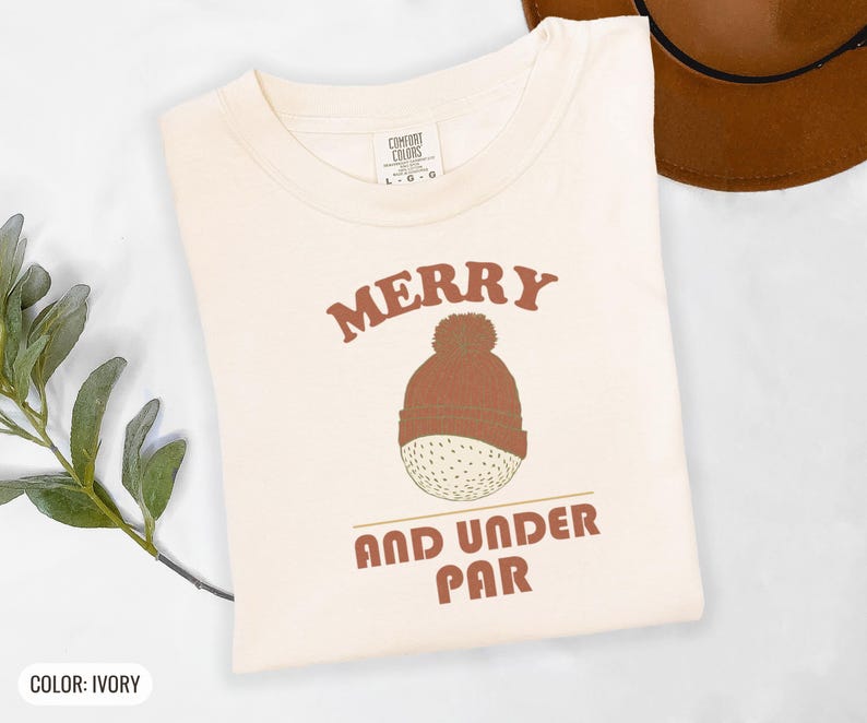 Ivory Comfort Colors t-shirt printed with the words Merry And Under Par and a drawing of a dimpled golf ball wearing a tiny red and white striped knit winter beanie with a green p, folded flat lay
