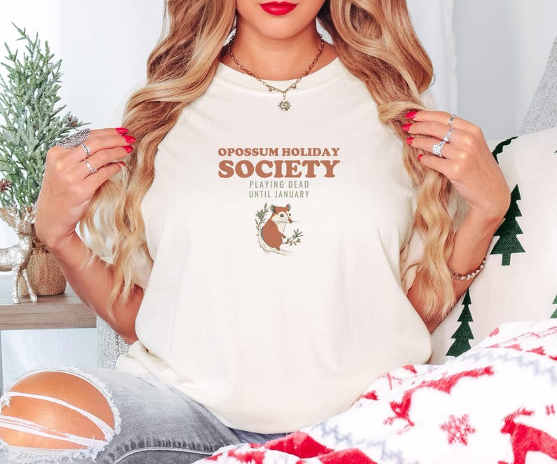 Ivory Comfort Colors t-shirt printed with the words Opossum Holiday Society Playing Dead Until January and a drawing of an opossum on a pine branch, worn by a woman