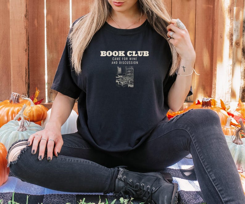 Black Comfort Colors t-shirt printed with the words Book Club Came For Wine And Discussion and a drawing of a cozy living room with books and wine glasses on a coffee table, worn by a woman