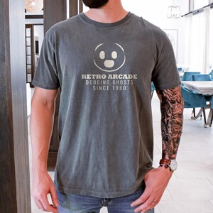 Pepper Comfort Colors t-shirt printed with the words Retro Arcade Dodging Ghosts Since 1980 and a drawing of a maze with a Pac-Man character, worn by a man
