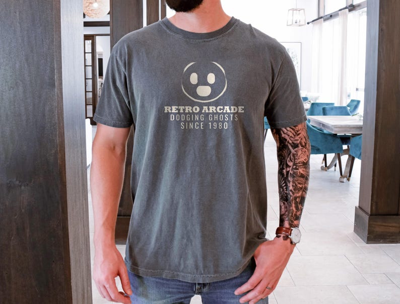 Pepper Comfort Colors t-shirt printed with the words Retro Arcade Dodging Ghosts Since 1980 and a drawing of a maze with a Pac-Man character, worn by a man