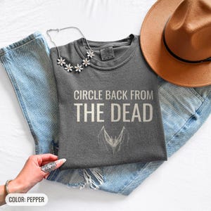 Pepper Comfort Colors t-shirt printed with the words Circle Back From The Dead and a drawing of a vintage silver paperclip bent into the shape of a bat, folded flat lay