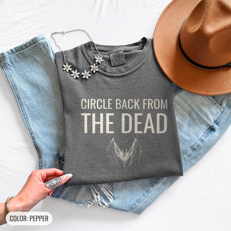 Pepper Comfort Colors t-shirt printed with the words Circle Back From The Dead and a drawing of a vintage silver paperclip bent into the shape of a bat, folded flat lay