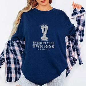 True Navy Comfort Colors t-shirt printed with the words Enter At Your Own Risk I Am Reading and a drawing of a brass candlestick with a single burning candle dripping wax, worn by a woman