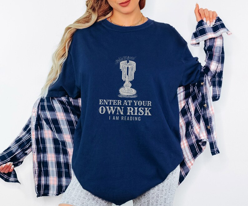 True Navy Comfort Colors t-shirt printed with the words Enter At Your Own Risk I Am Reading and a drawing of a brass candlestick with a single burning candle dripping wax, worn by a woman