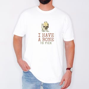 White Comfort Colors t-shirt printed with the words I Have A Bone To Pick and a drawing of an anatomical etching of a corvid raven skull entwined with dried wild thistle blossoms, worn by a man