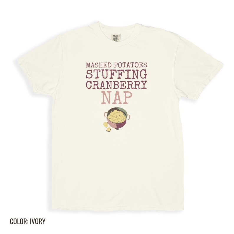 Ivory Comfort Colors t-shirt printed with the words Mashed Potatoes Stuffing Cranberry Nap and a drawing of a heavy enamel-coated cast iron Dutch oven filled with creamy mashed potatoes, steam lines, folded flat lay