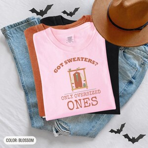 Blossom Comfort Colors t-shirt printed with the words Got Sweaters? Only Oversized Ones and a drawing of a knitted wool cardigan hanging from a simple wooden clothes hanger, worn by a woman