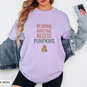 Orchid Comfort Colors t-shirt printed with the words Reading Writing Recess Pumpkins and a drawing of a stack of three vintage schoolbooks with a small orange pumpkin sitting on top, worn by a woman