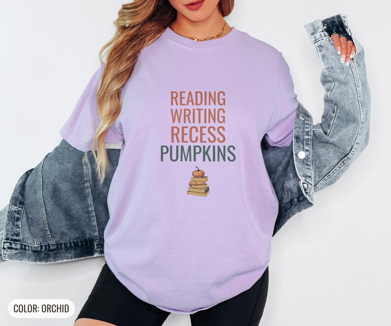 Orchid Comfort Colors t-shirt printed with the words Reading Writing Recess Pumpkins and a drawing of a stack of three vintage schoolbooks with a small orange pumpkin sitting on top, worn by a woman