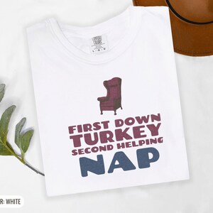 White Comfort Colors t-shirt printed with the words First Down Turkey Second Helping Nap and a drawing of a vintage brown recliner with a footrest extended outward, folded flat lay