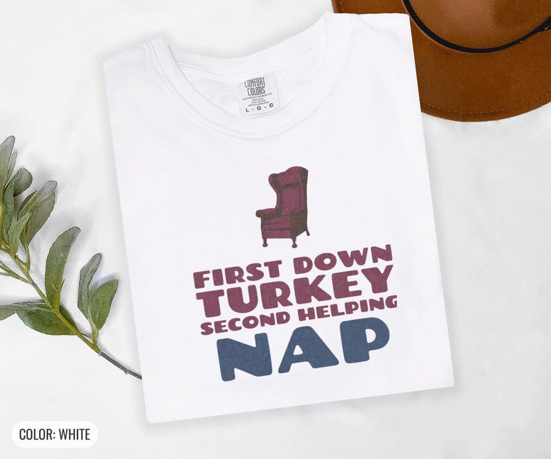 White Comfort Colors t-shirt printed with the words First Down Turkey Second Helping Nap and a drawing of a vintage brown recliner with a footrest extended outward, folded flat lay